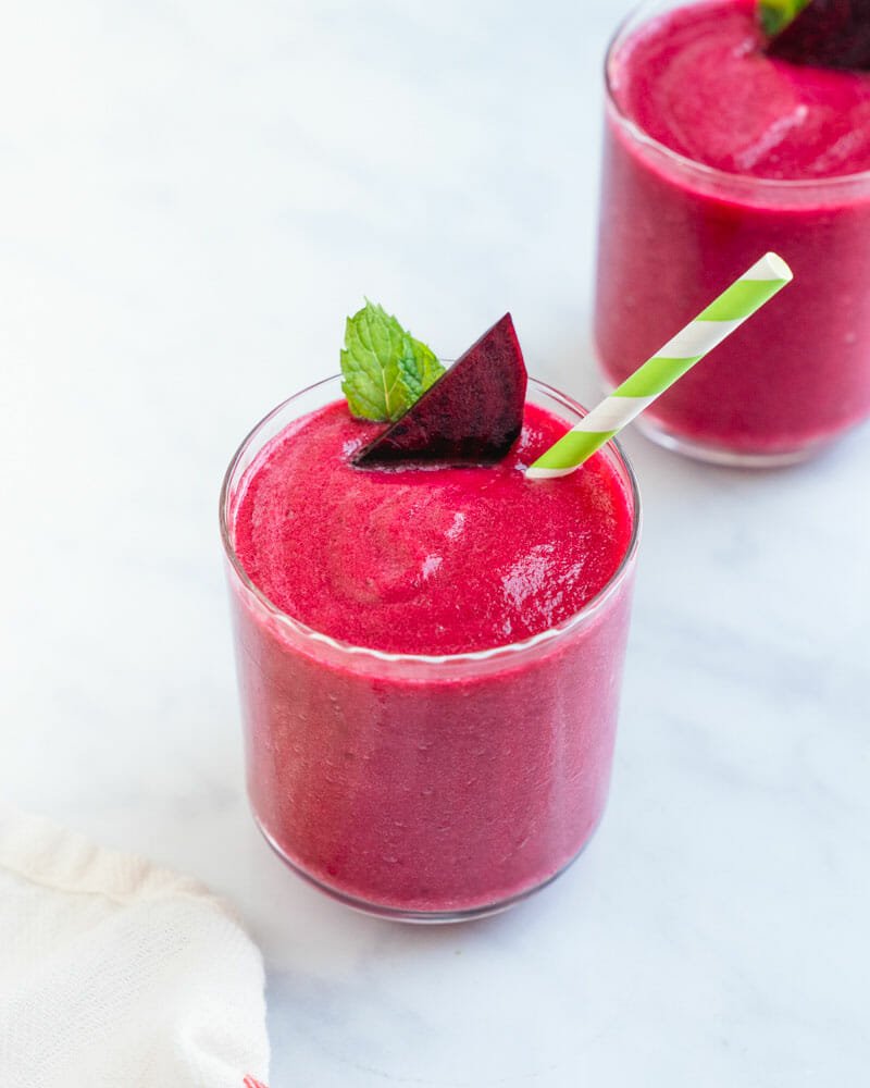 Two dark pink smoothie drinks, one with a green and white striped straw, a slice of beet and a leaf.