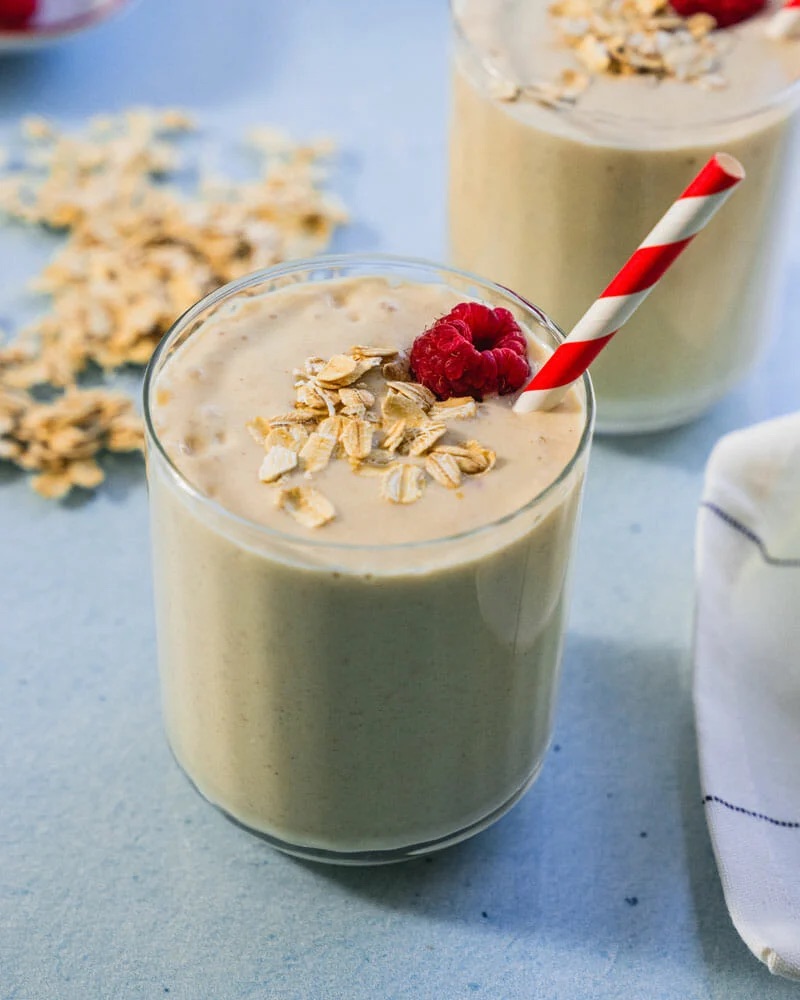Pale brown oatmeal smoothie in a glass with oats on top and striped red and white straw.