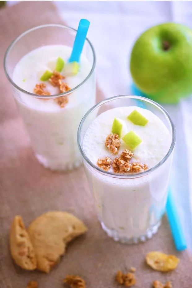 White frothy smoothie in a glass with pieces of apple and granola on top.