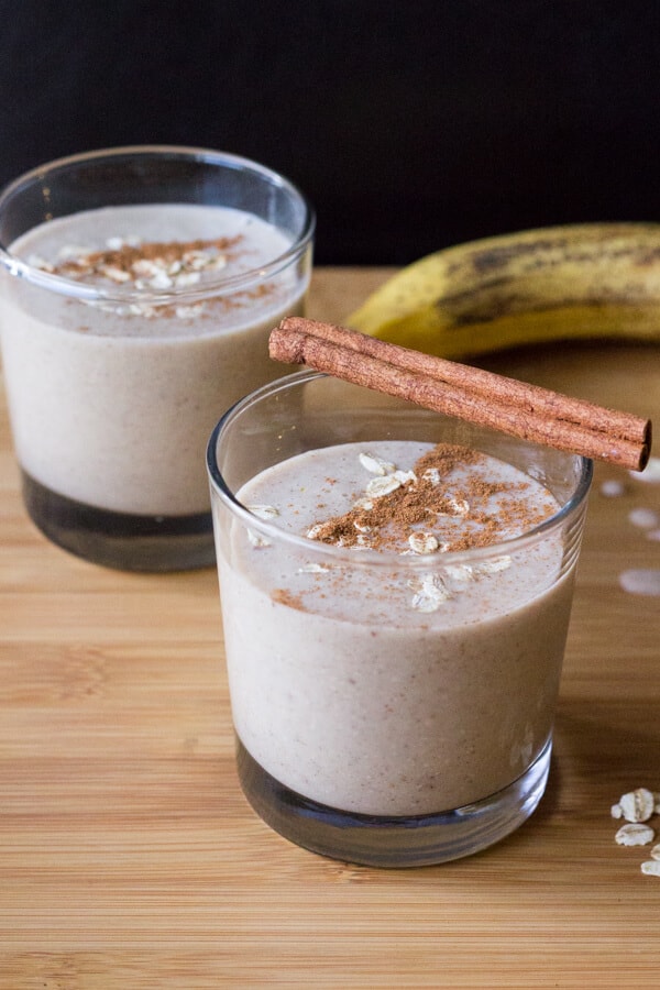 Two glasses of pale brown oatmeal smoothie with cinammon sticks balancing on one glass.