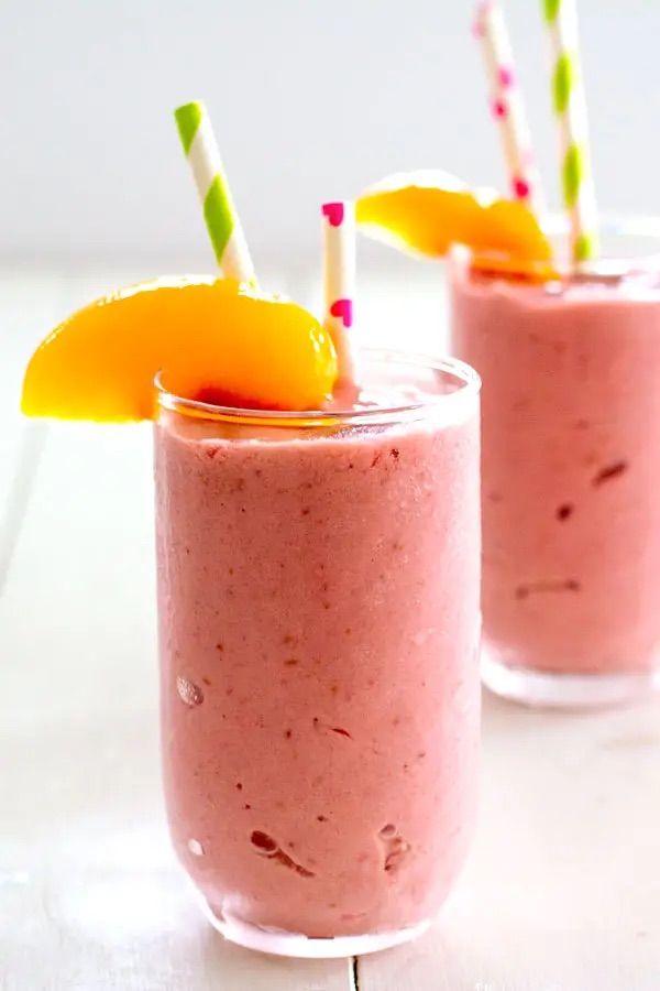 Two glasses of pink smoothie each with two striped straws and a slice of peach.