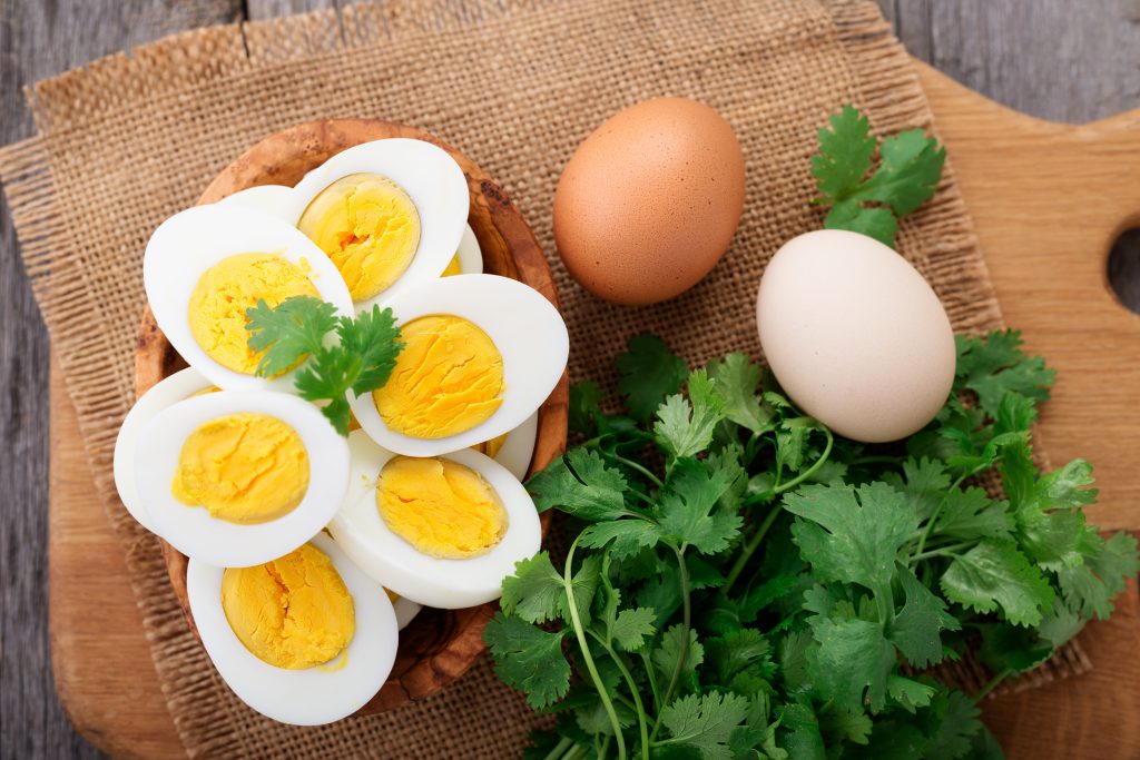 Wooden bowl of halved hard boiled eggs, and two whole eggs on a canvas background and wooden chopping board with green leaves..