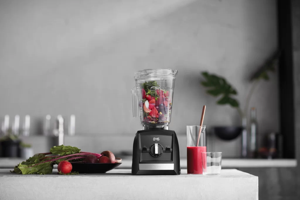 Black blender with digital display in a kitchen. Jar is full of fruit and vegetables ready to blend. Blender is next to a glass half-full of a red drink.