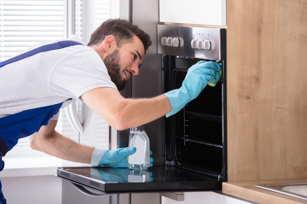 Man with beard wearing rubber gloves and overalls cleaning an oven