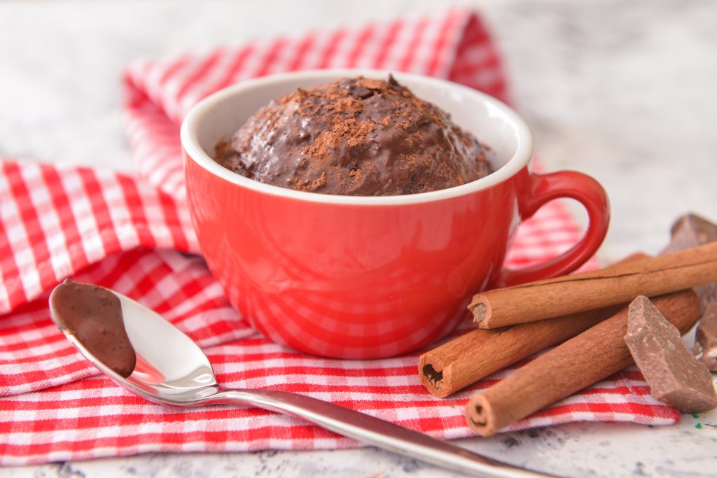 Chocolate Mug Cake in a red mug with cinnamon sticks and spoon by side, on checked napkin