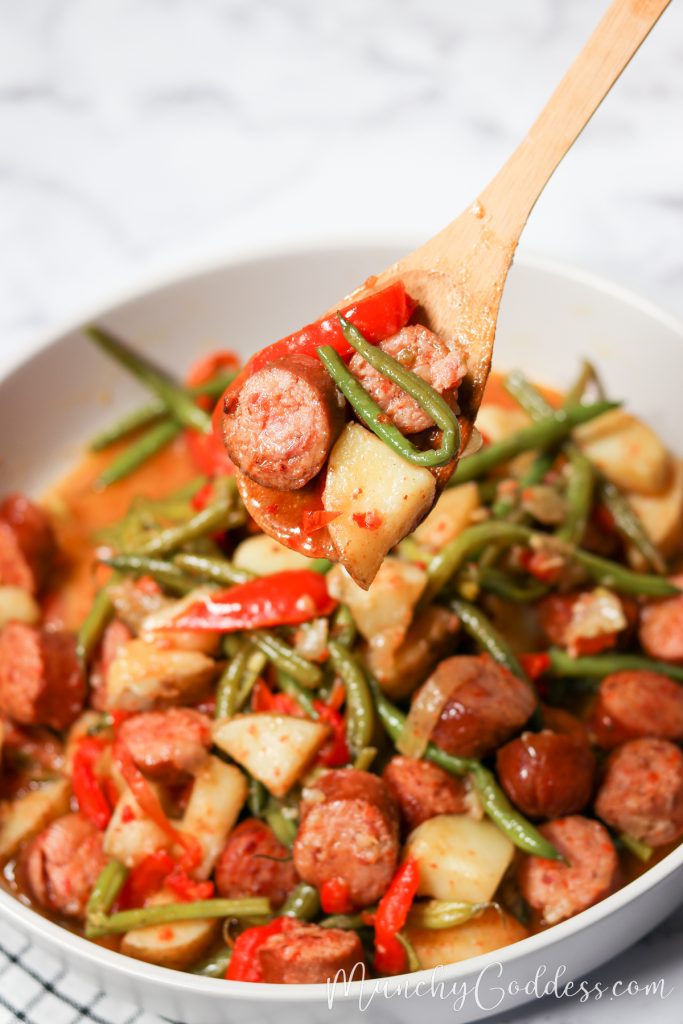 Green beans with sausage and red peppers in a white dish and scooped in a wooden spoon