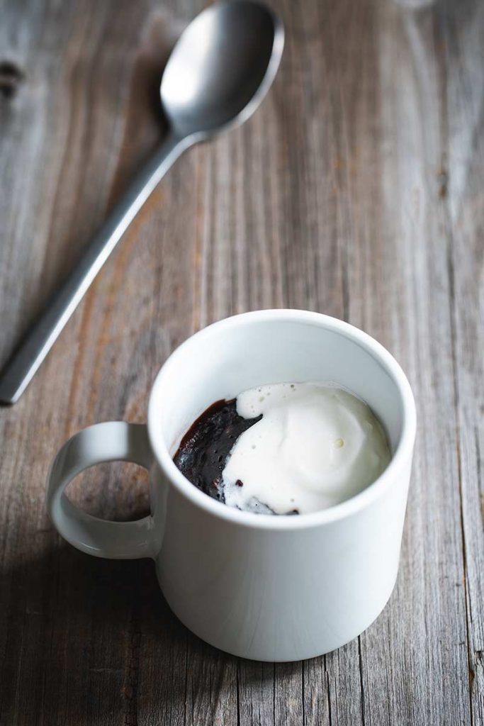 Chocolate brownie cooked in a mug, with cream on top and spoon lying on table