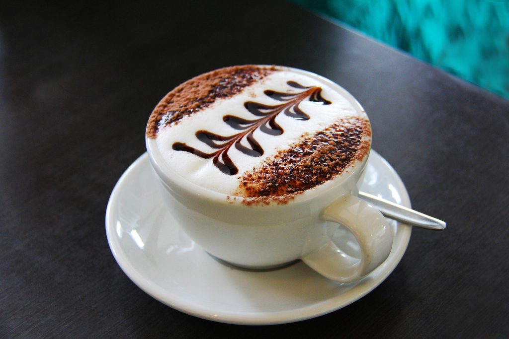 Coffee in a white cup with chocolate powder sprinkled on top and a design in chocolate sauce on white milk foam