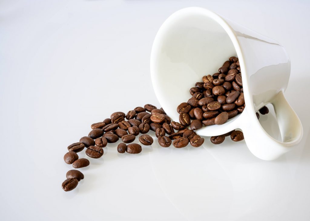 White cup on its side with coffee beans spilling out onto white background