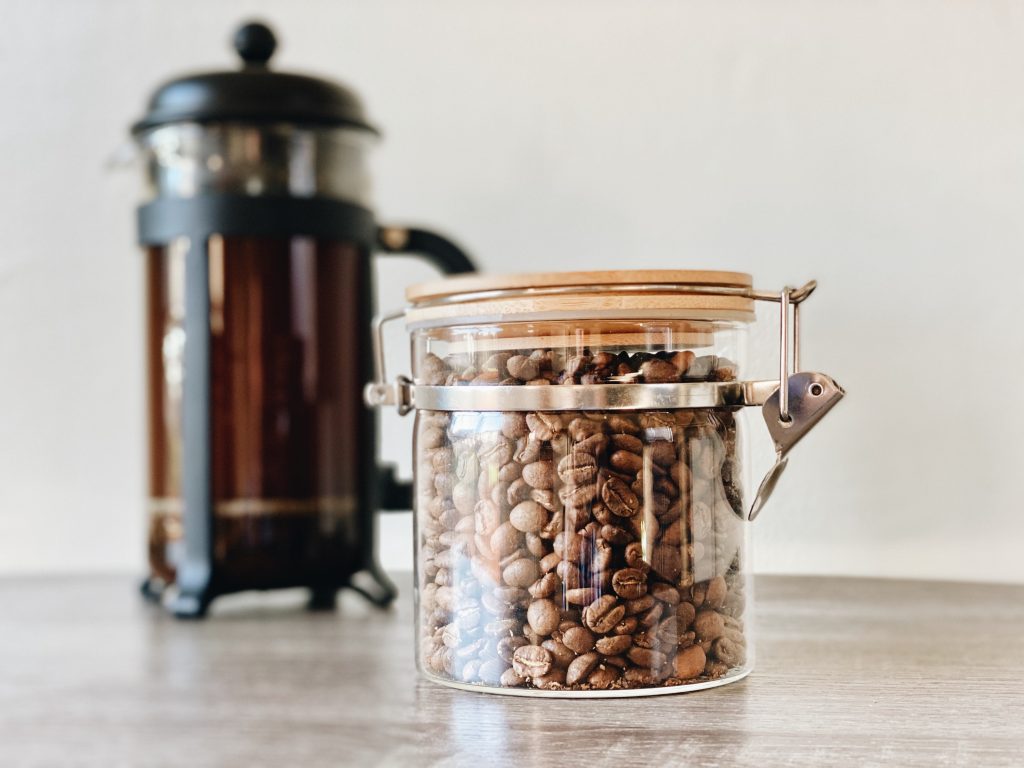 French press in background with jar of coffee beans in foreground