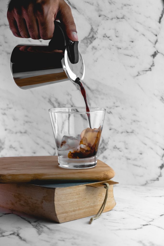 Glass with ice cubes balancin on book and chopping board, with black coffee being poured into it